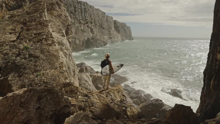 Person holding a surfboard watching the sea among rocks at the edge of a cliff