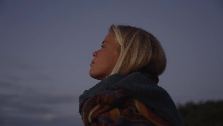 A persons head and shoulders are shown in profile the person is wrapped in a patterned blanket the person looks up toward the sky the background is a flat expanse of grayblue and the scene is outdoors in low light