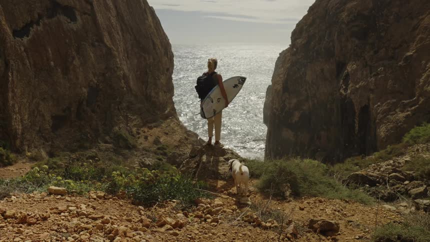 Female surfer carrying her board and her dog hiking to cliffs overlooking the coast