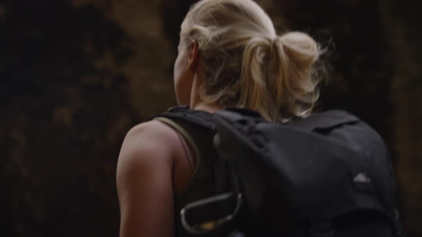 Blondehaired woman with bun carries black backpack in rocky setting with natural lighting
