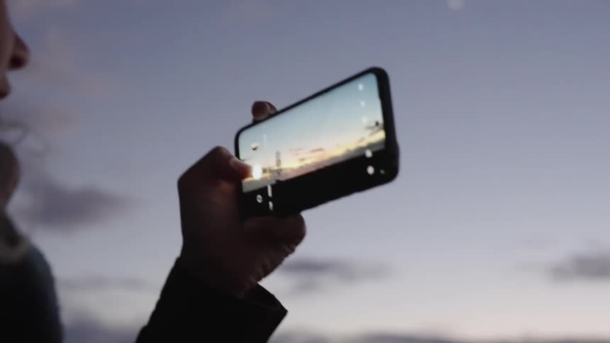 The video is a closeup profile shot of a young woman standing outdoors at dusk looking down at the sunset captured on her smartphone screen her blonde hair frames her face and the sky behind her is cloudy and fading to dark