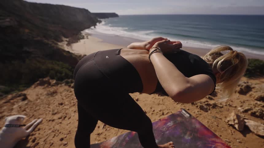 A person stands on a mat the person bends forward and locks their hands behind their back the ground is dirt and rock the ocean and a beach are visible below the cliff and a dog lies on the ground nearby