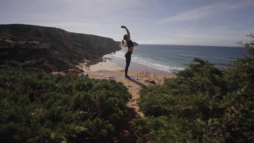 Woman practices yoga on top of a mountain overlooking the coast below