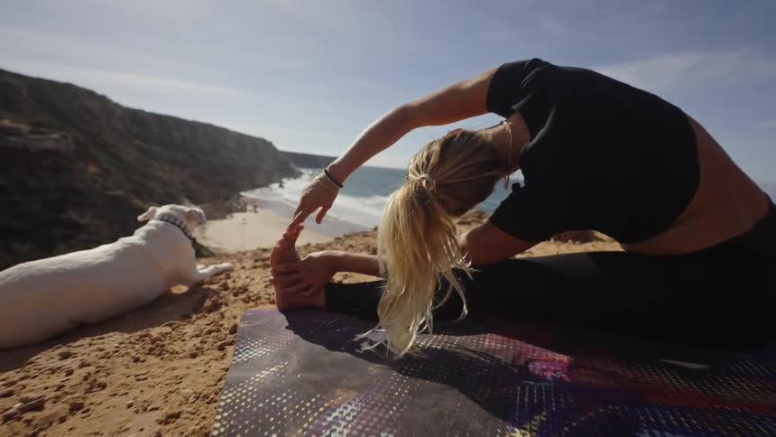 Woman practices yoga on a cliff with a white dog nearby