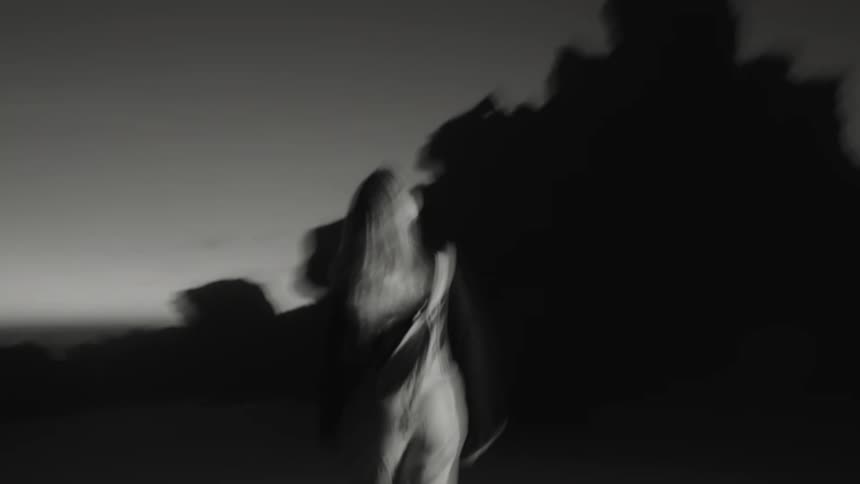 The video is a black and white shot of a person with blonde hair walking quickly away from the camera in a lowlight or blurred environment they are wearing a dark top and what appears to be a white crossbody bag or sling with the background co