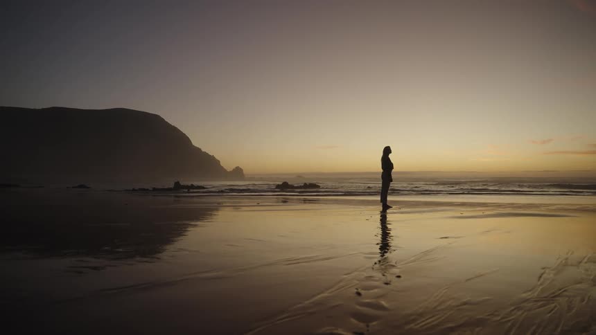 Person standing on wet sand watching the sea at sunset