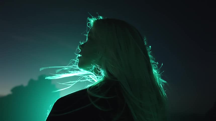 The outline of a persons head and shoulders is visible long hair moves due to air current bright green light illuminates the hair and outline from behind the setting is outdoors during darkness