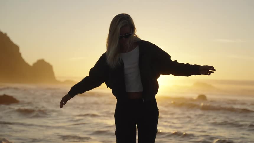 A person stands on a beach the persons arms are outstretched and she moves her body water from waves washes around their feet the sun creates a glow behind the person and cliffs are visible in the background