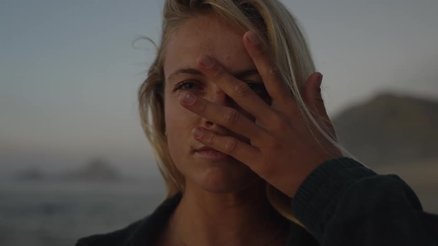 A persons face is shown in a closeup one hand covers the right side of the face the person looks directly forward the background is a blurry gray beach and water and the scene is outdoors in low light