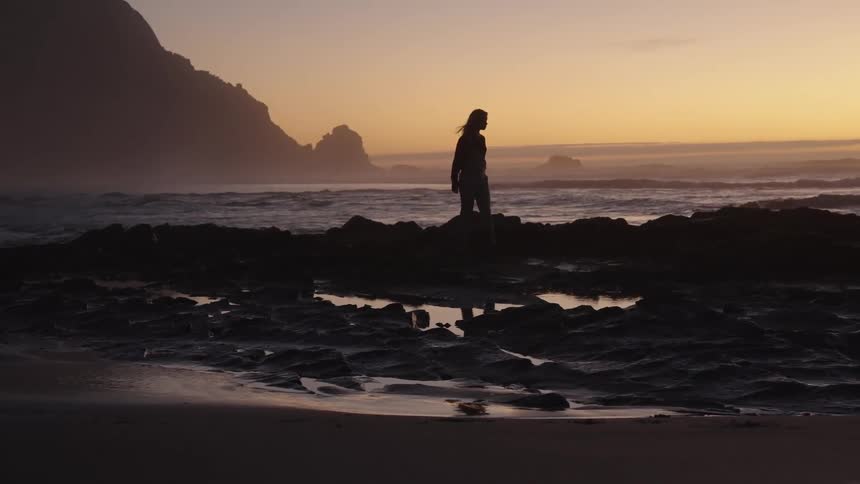 Silhouette of a female walking on rocky shoreline at the beach at dusk