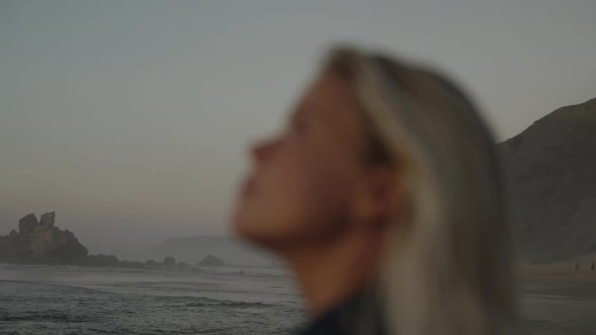 A blonde woman stands on the shoreline gazing at the sky during sunset