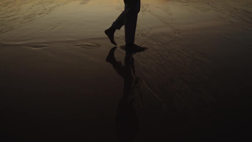 A low angle close view captures the bare feet of a person wearing dark pants walking on the wet reflective sand of a beach with the bright sunset light reflecting in the foreground