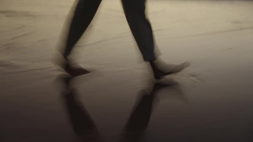 A closeup shot focuses on the bare legs of a person walking across wet dark sand the water from the waves is washing over their feet and ankles as they step
