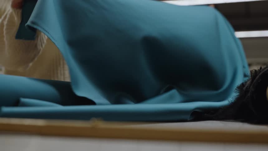 A close view shows the hands of a woman wearing a white knitted sweater carefully unrolling and smoothly running over a piece of teal or dark blue fabric on a wooden cutting table in a bright sewing studio or textile factory