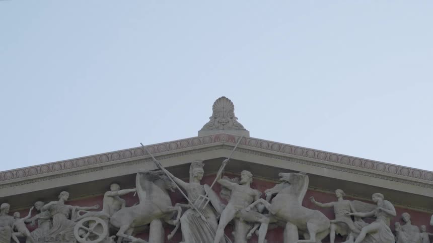 A medium view of the triangular pediment of a large greek temple structure is filled with detailed stone sculptures of horses and warriors beneath a pale blue sky