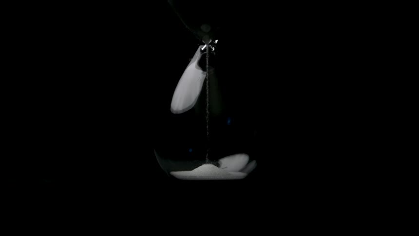 A glass bulb hangs suspended from a string against a solid black background white sand flows downward from the upper section to the lower section and the white material piles up in the bottom curve of the container