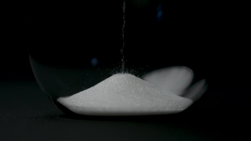 A close dramatic view shows a thin stream of white sand falling from the upper bulb into a growing pile in the bottom bulb of a glass hourglass against a solid black background