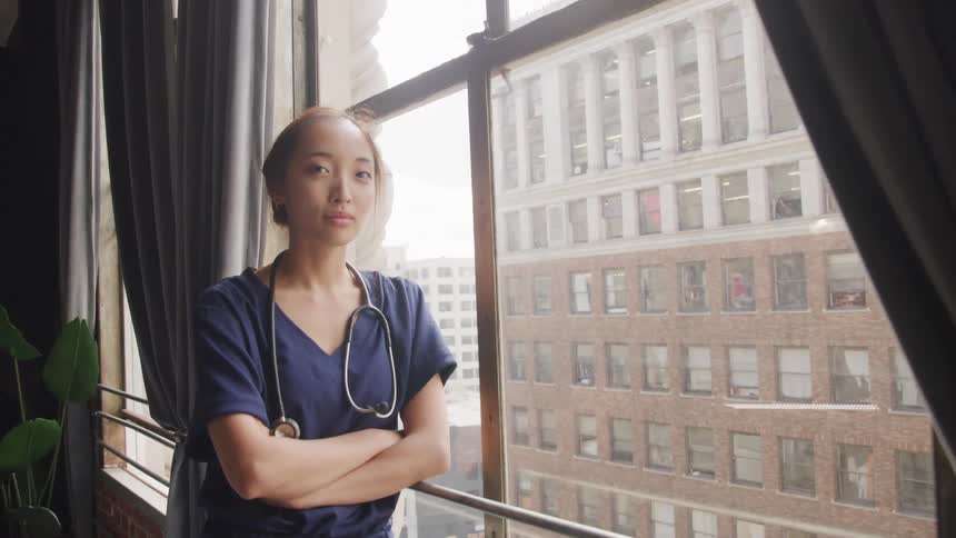 A nurse rests against a window at midday a stethoscope draped over her shoulder with a highrise building visible outside