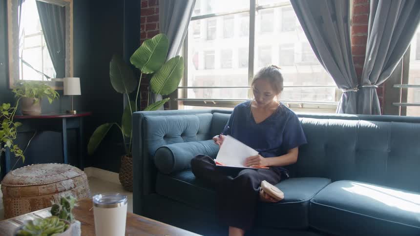 A woman writes in a notebook on a blue sofa in a sunlit living room