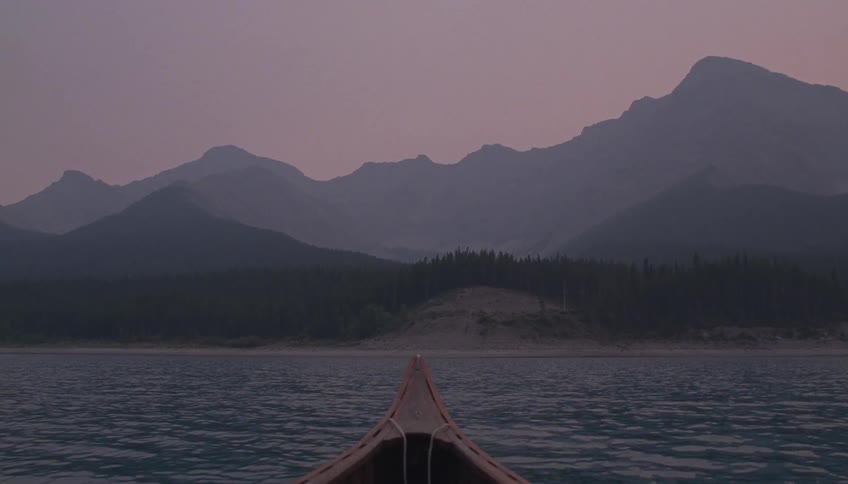 Canoe on a large body of water approaches the shoreline with large layered mountains rising up in the background under a dim hazy sky