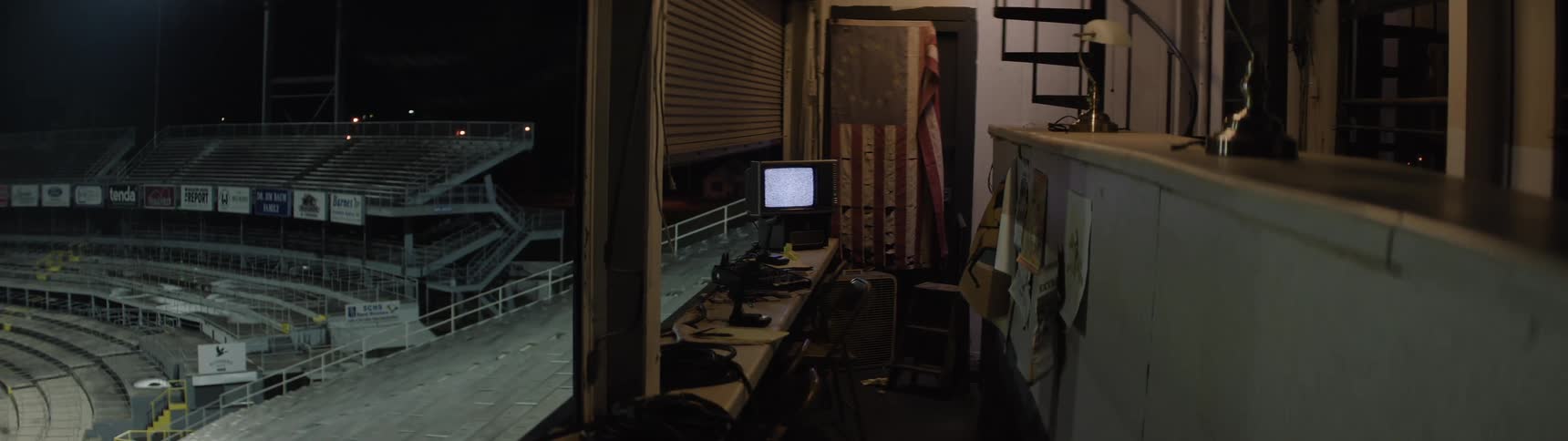 A dim press box room overlooks a lit stadium with a small monitor and scattered gear lining a long counter by the window
