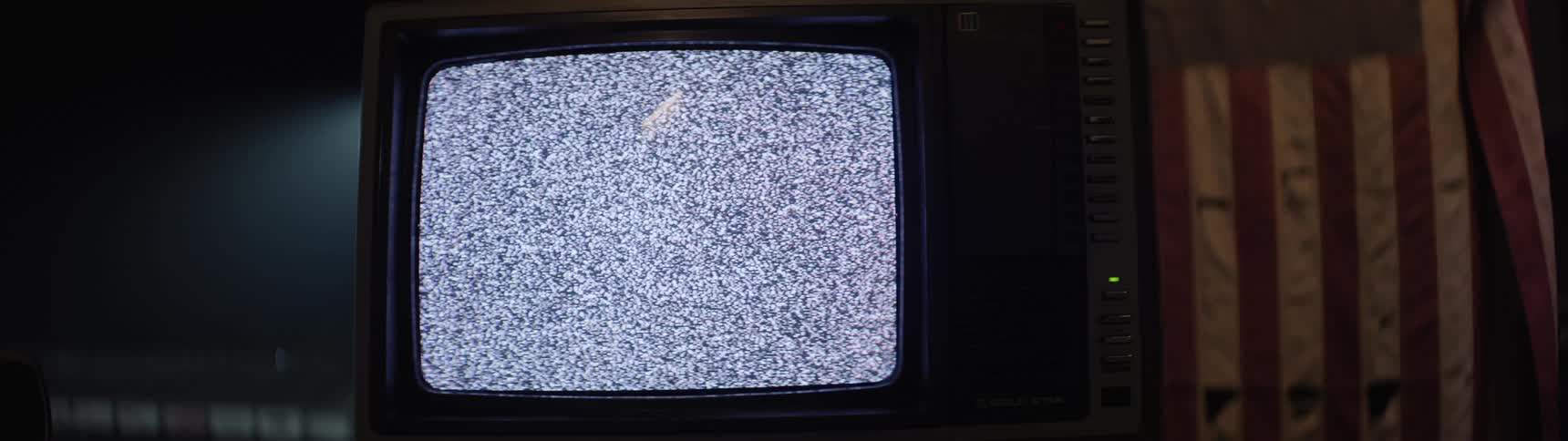 A vintage crt television shows static in a dim room beside a worn american flag
