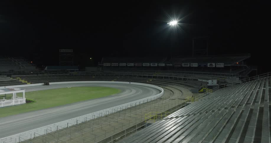 A night racetrack curves through empty grandstands under a single bright floodlight