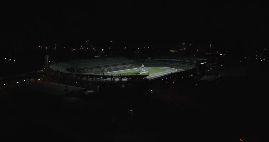 The video is a wide highangle shot of an empty ovalshaped racetrack stadium at night dimly lit by stadium lights the grandstands are dark against the black sky while the bright lights focus mainly on the grass infield and the dirt track
