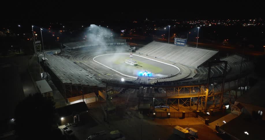 An aerial view shows a large oval racetrack stadium at night bright field lights illuminate the empty track surface people are gathered in the field part and steam rises from the center track area