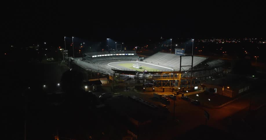 The video is a wide aerial shot of a speedway or dirt track stadium lit up at night the surrounding area is dark with scattered city lights visible in the background