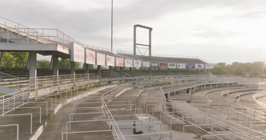 The video is an exterior shot of a large curved concrete stadium with tiered seating and metal railings appearing to be for a racetrack or outdoor arena the stands are empty and look slightly worn with a line of sponsor signs running along the