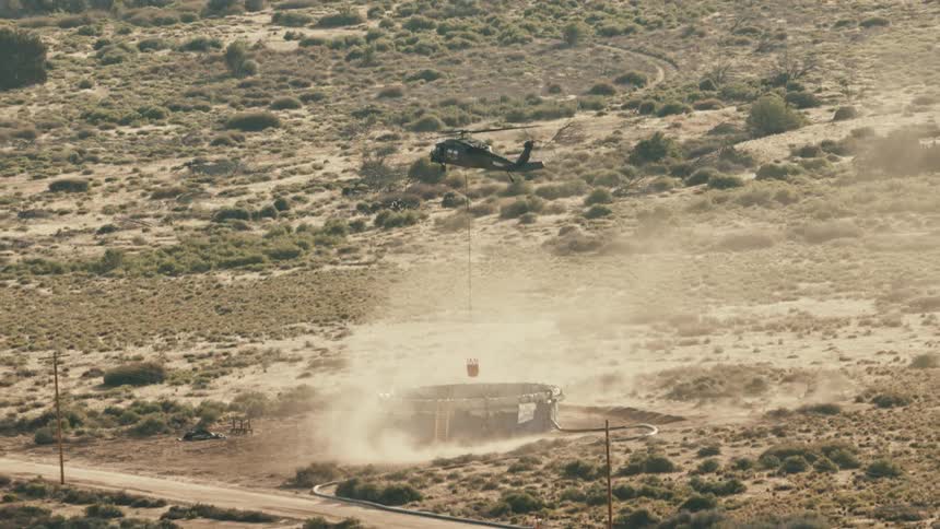 The video is an aerial shot showing a darkcolored helicopter lifting a bucket from a portable water tank on the ground kicking up dust in a dry scrubcovered landscape this suggests the helicopter is performing a watercollection mission