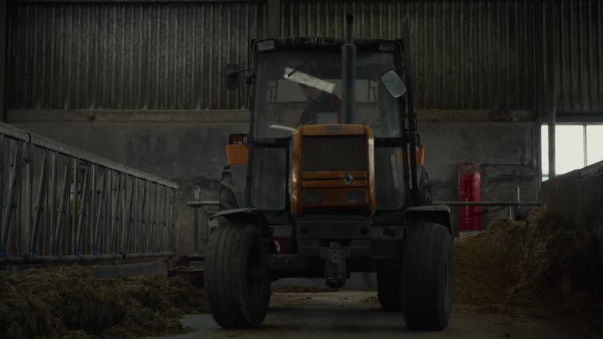 A yellow and black tractor with a driver is seen moving inside a large dimly lit barn the vehicle is centered between dark piles of feed and the corrugated metal walls of the structure are visible