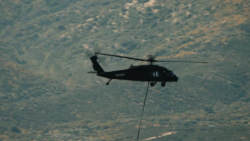 A black helicopter flies horizontally against a background of dry hills and mountains