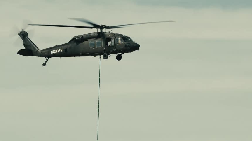 A low angle medium view shows a dark gray black hawk helicopter flying across a pale cloudy sky with a long green rescue rope dangling below it
