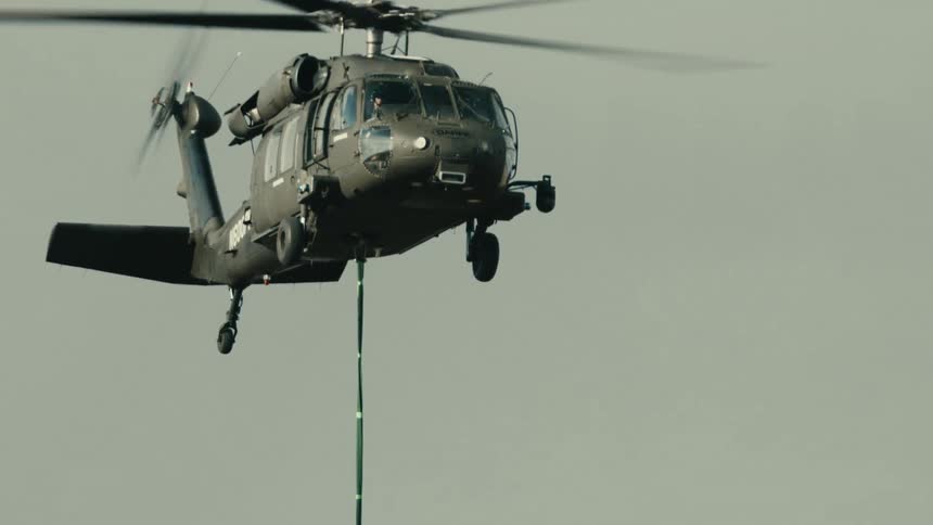 A low angle close view shows a dark military helicopter in flight with its rotor blades spinning and a long green rescue rope dangling below against a pale overcast sky