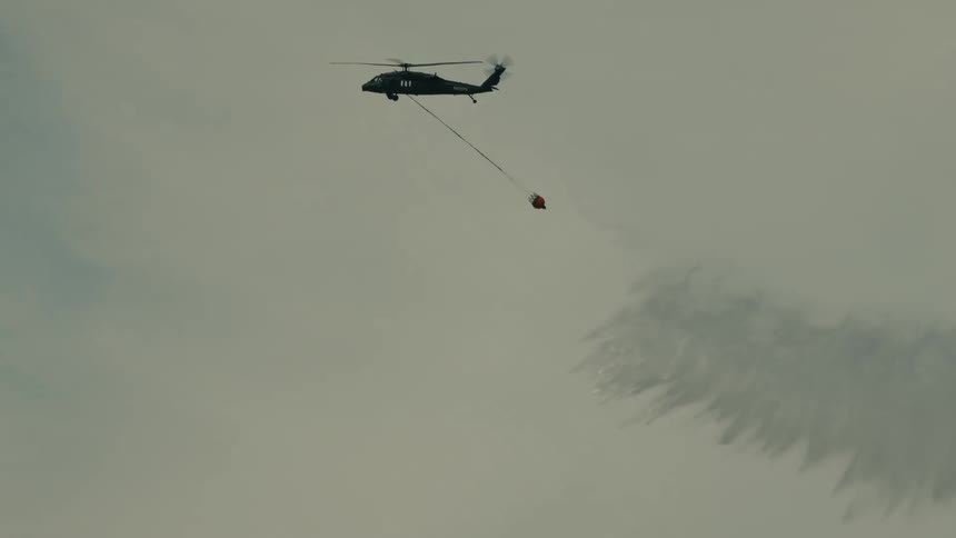 The video is an extreme closeup shot of a black hawk helicopter silhouetted against a pale sky carrying a bucket on a long line the helicopter is performing a water drop with a large white spray of water visible below it
