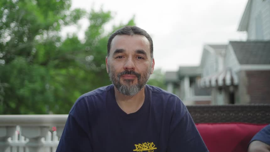 A medium close view of a man with dark hair and a gray beard wearing a dark blue tshirt with a yellow graphic as he sits on a red cushion outdoors and smiles faintly at the camera