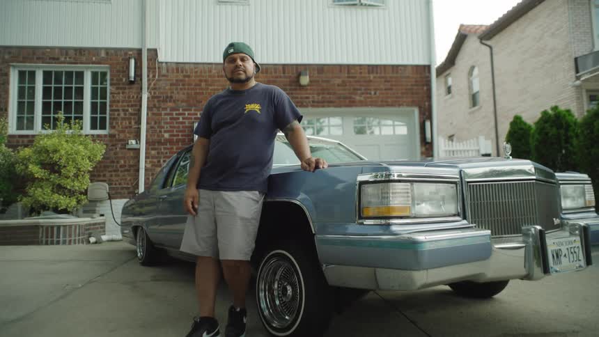 A man stands outside leaning against the front fender of a large blue classic american sedan he wears a baseball cap and casual clothes and the background shows a driveway and brick residential buildings on a clear day