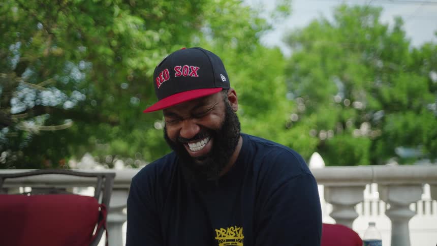 A man with a full beard and a baseball cap sits outdoors he laughs heartily and widely at something offcamera the background consists of lush green trees and a white decorative railing illuminated by bright daylight