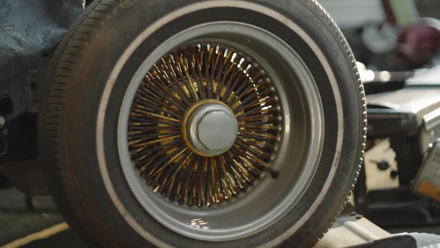 A vintage wirespoke wheel with a whitewall tire sits on a classic car chassis in warm sunlight