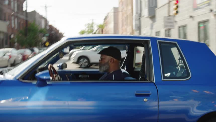 A middle aged black man driving a blue vintage vehicle on a suburban street during daytime