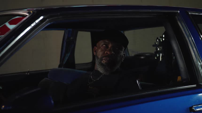 The video is an interior shot of a man sitting in the drivers seat of a glossy blue classic car he is wearing a dark cap and a silver chain and briefly looks directly toward the camera