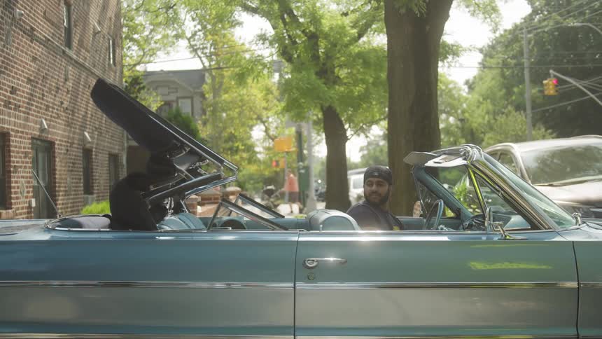A side medium view of a man with a beard wearing a dark shirt and baseball cap as he smiles while sitting inside a classic light blue convertible with the roof lowering on a tree lined street
