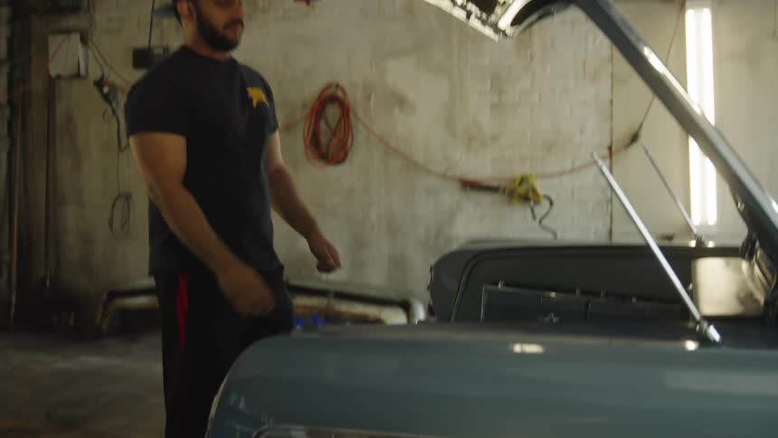 A man wearing a dark shirt and baseball cap opens the trunk of a classic car inside a dimly lit garage or workshop the camera then focuses on the custom contents of the trunk which include chrome hydraulic pumps and a large speaker for a lowride