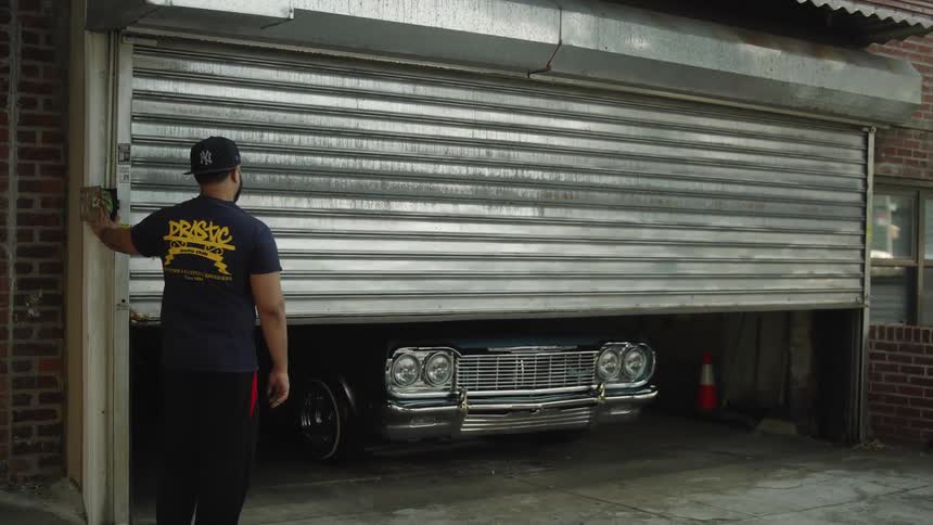 Man wearing a blue cap operating a metal garage door partially open with a classic car inside