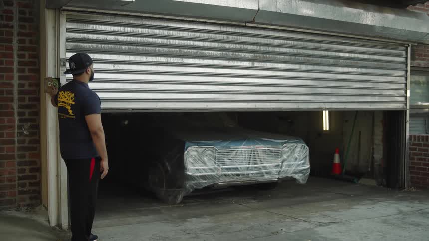 A black man wearing a hat presses a button at midday opening a garage to reveal a car inside
