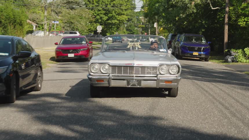 A man sits in an openroof car driving down a sunny street lined with parked cars