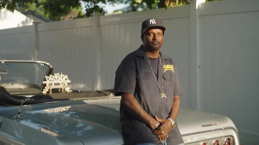 A medium view shows a man with a mustache and goatee wearing a dark work shirt with a yellow logo a yankees cap and gold chains as he stands leaning on a vintage light blue and silver convertible car on a sunny day