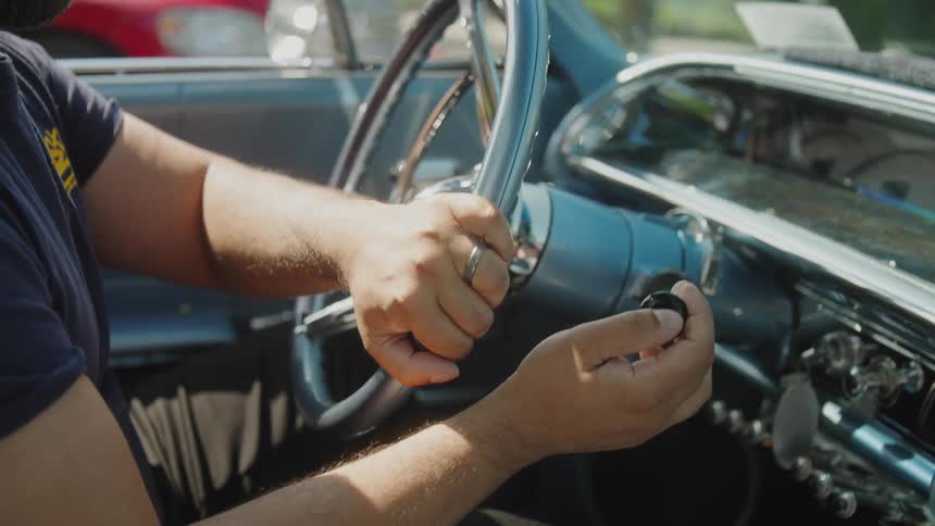 A close view of a bearded man wearing a dark shirt in a classic convertible car holding the steering wheel and inserting the key into the ignition on a sunny day